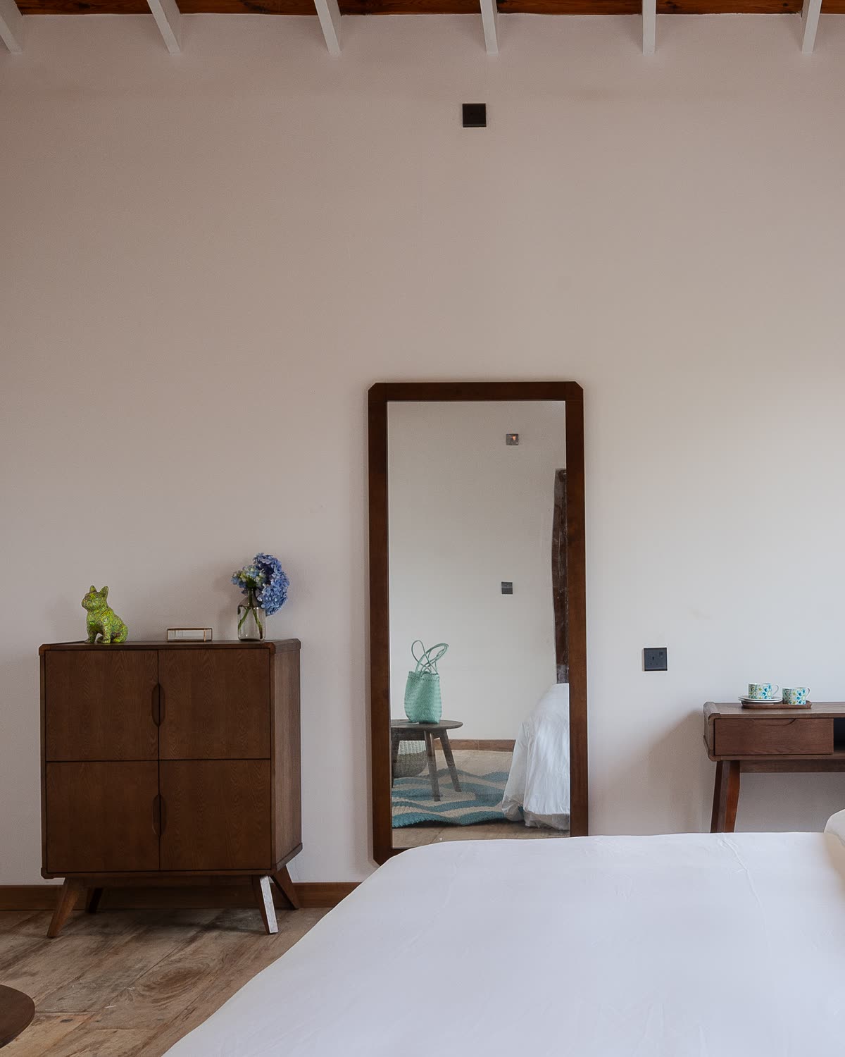 A chalet bedroom: a walnut sideboard with hydrangeas beside a tall floor mirror, a writing desk and white bed linen in the foreground