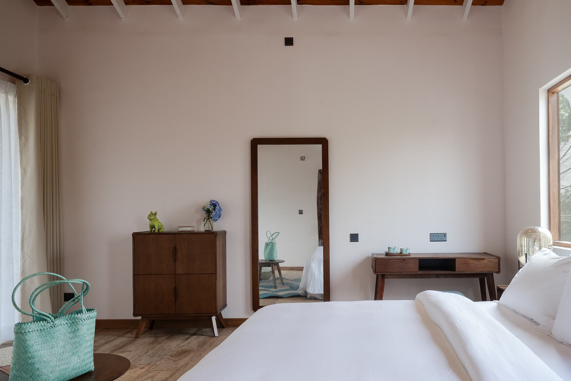 A chalet bedroom with a timber-joist ceiling: a walnut sideboard with hydrangeas, a tall floor mirror, a writing desk and white bed linen in soft window light