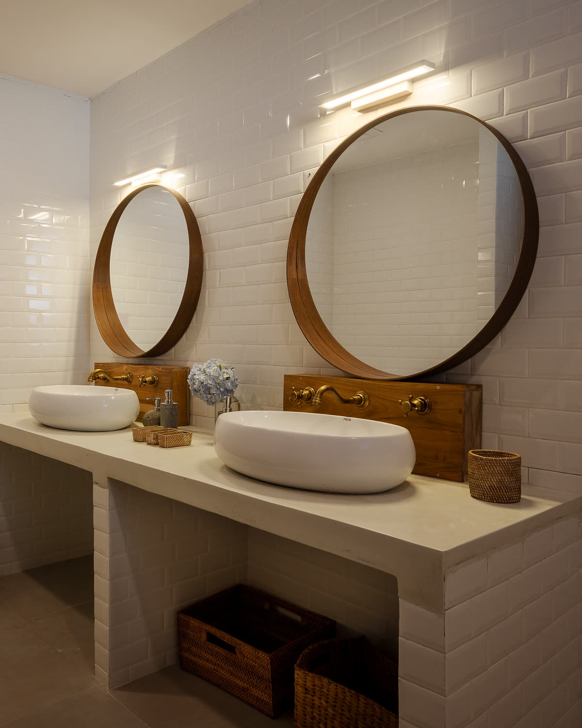 A double vanity in a chalet bathroom: two white basins, brass taps set in teak and round teak-framed mirrors against white tiles, woven baskets beneath