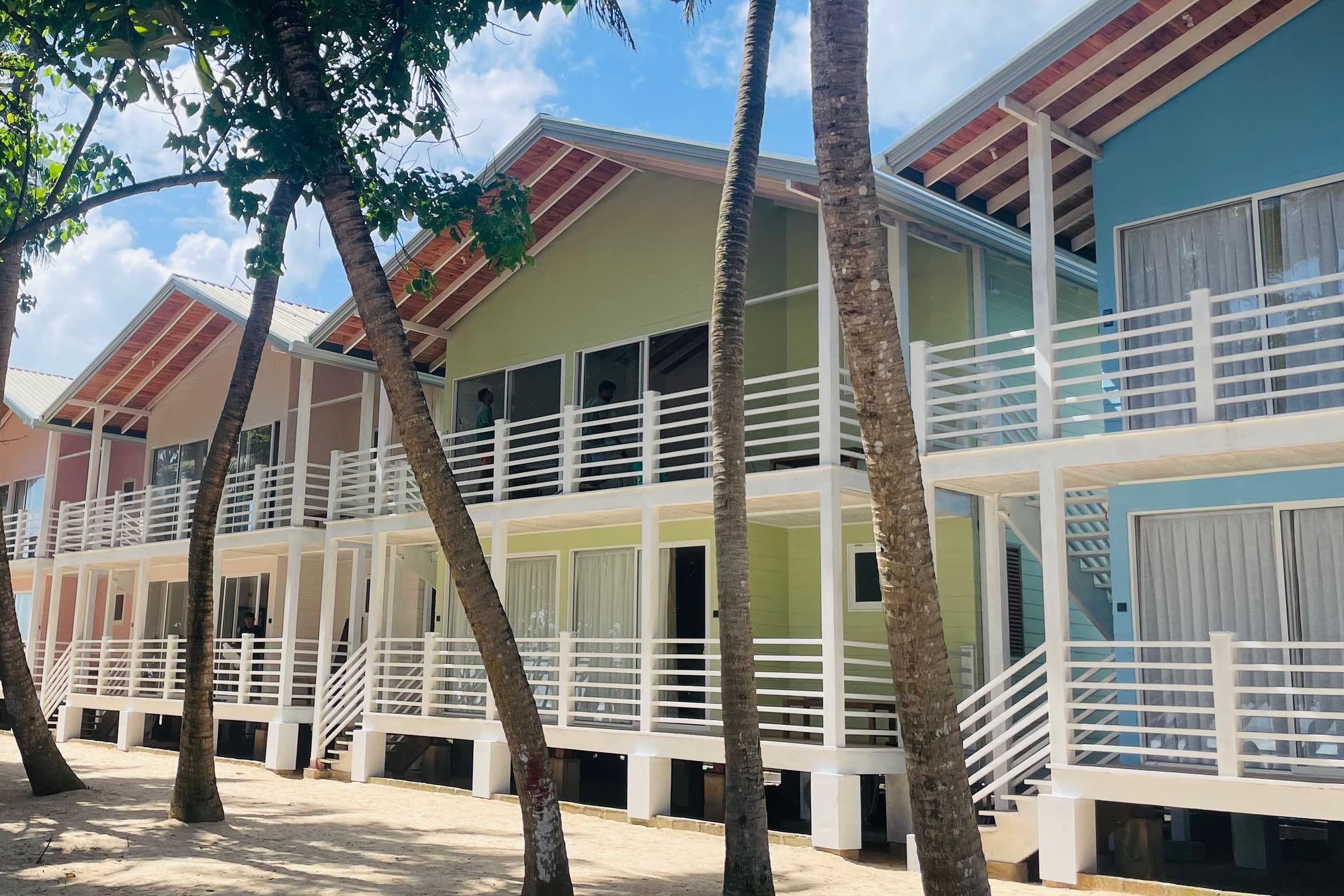 A row of two-storey timber chalets in pink, peach, green and blue with white balconies, seen between palm trunks on the sand under a blue sky