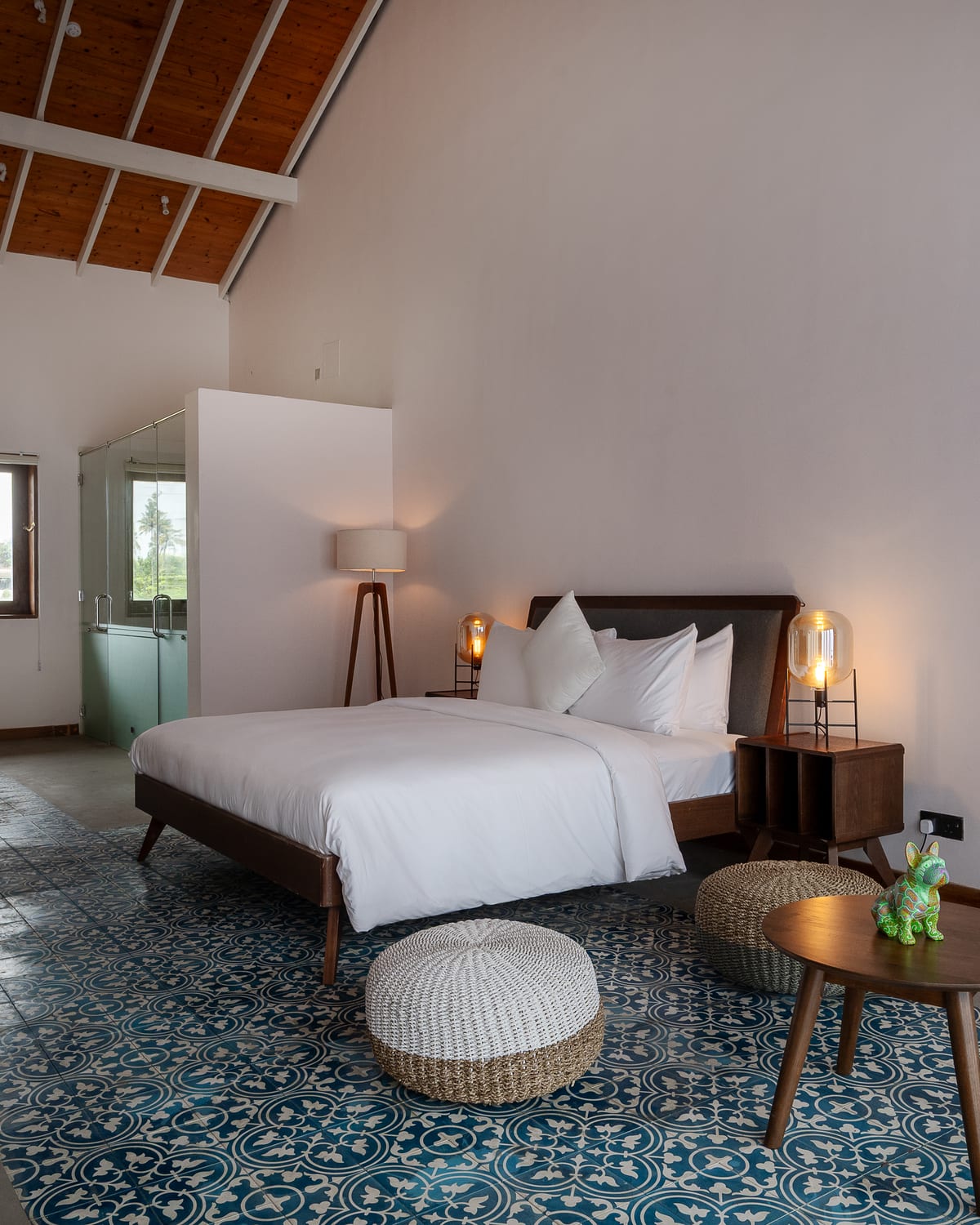 The Club Premier under a pitched timber ceiling: a king-size bed in white linen, blue and white patterned floor tiles, a woven pouf and a glass-walled shower beyond