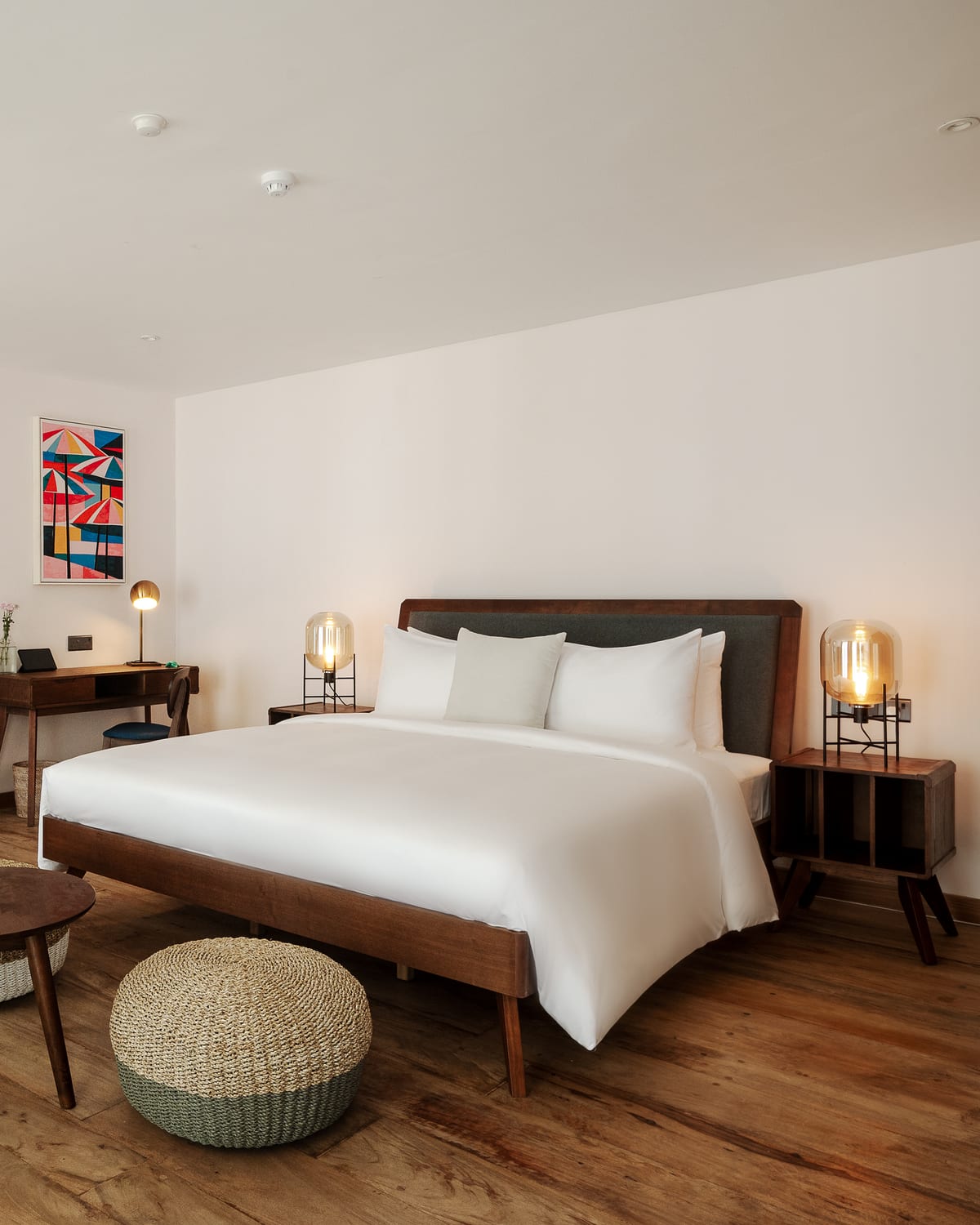 A Deluxe Chalet bedroom on the ground floor: a king-size bed in white linen with a timber frame, glass lamps on the side tables, a writing desk under a bright painting and woven poufs on a timber floor