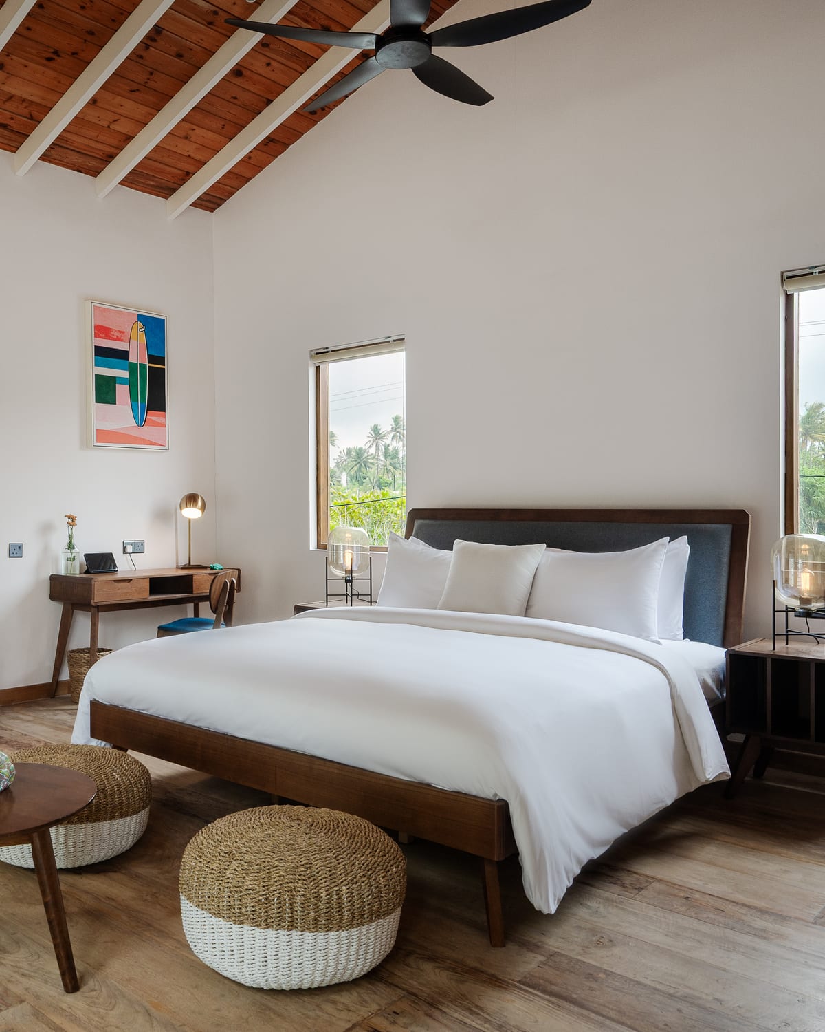 A Premier Chalet bedroom under a pitched timber ceiling with a fan: a king-size bed in white linen, a writing desk under a surfboard painting and a window onto palms