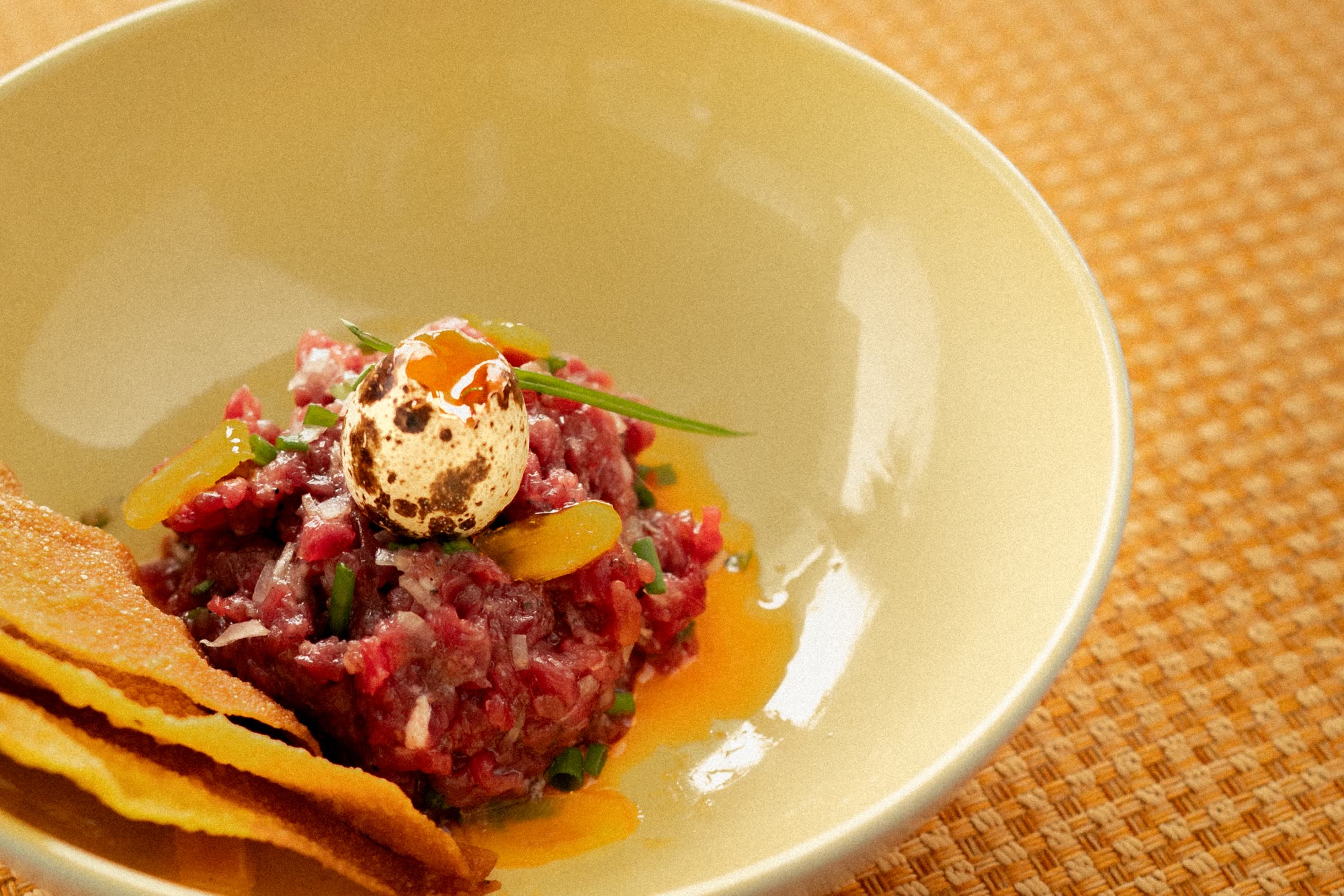 Beef tartare topped with a quail egg, with golden crisps, in a cream bowl on a woven orange placemat
