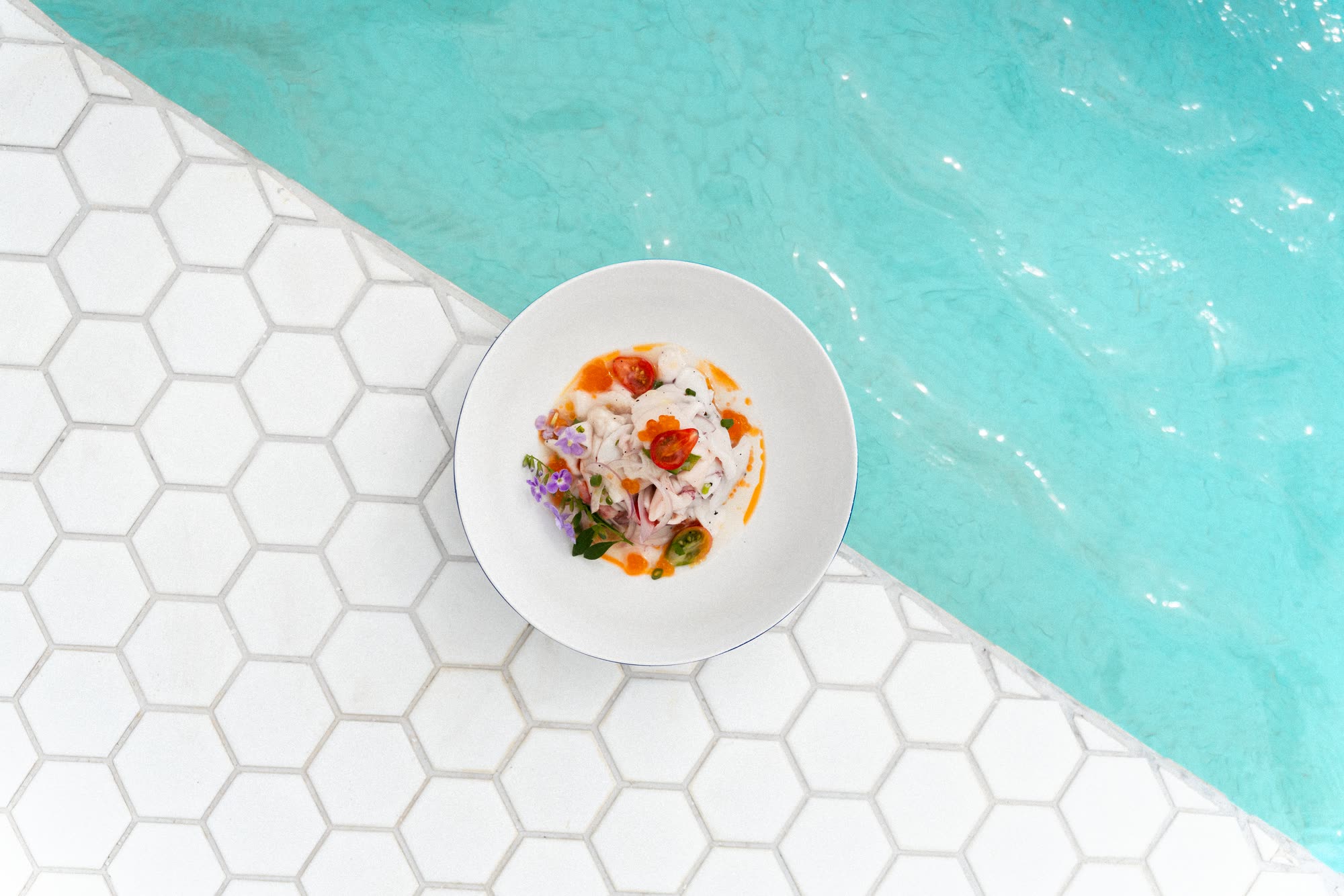 A white bowl of ceviche with cherry tomatoes, chilli and purple flowers, seen from above on white hexagonal tiles at the edge of the pool