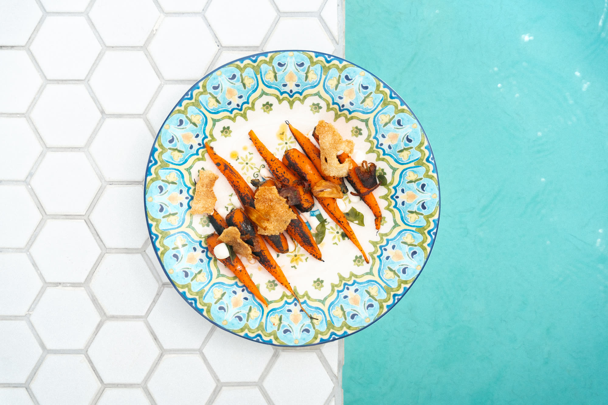 Charred carrots with crisps and herbs on a blue and green patterned plate, on white hexagonal tiles at the edge of the pool