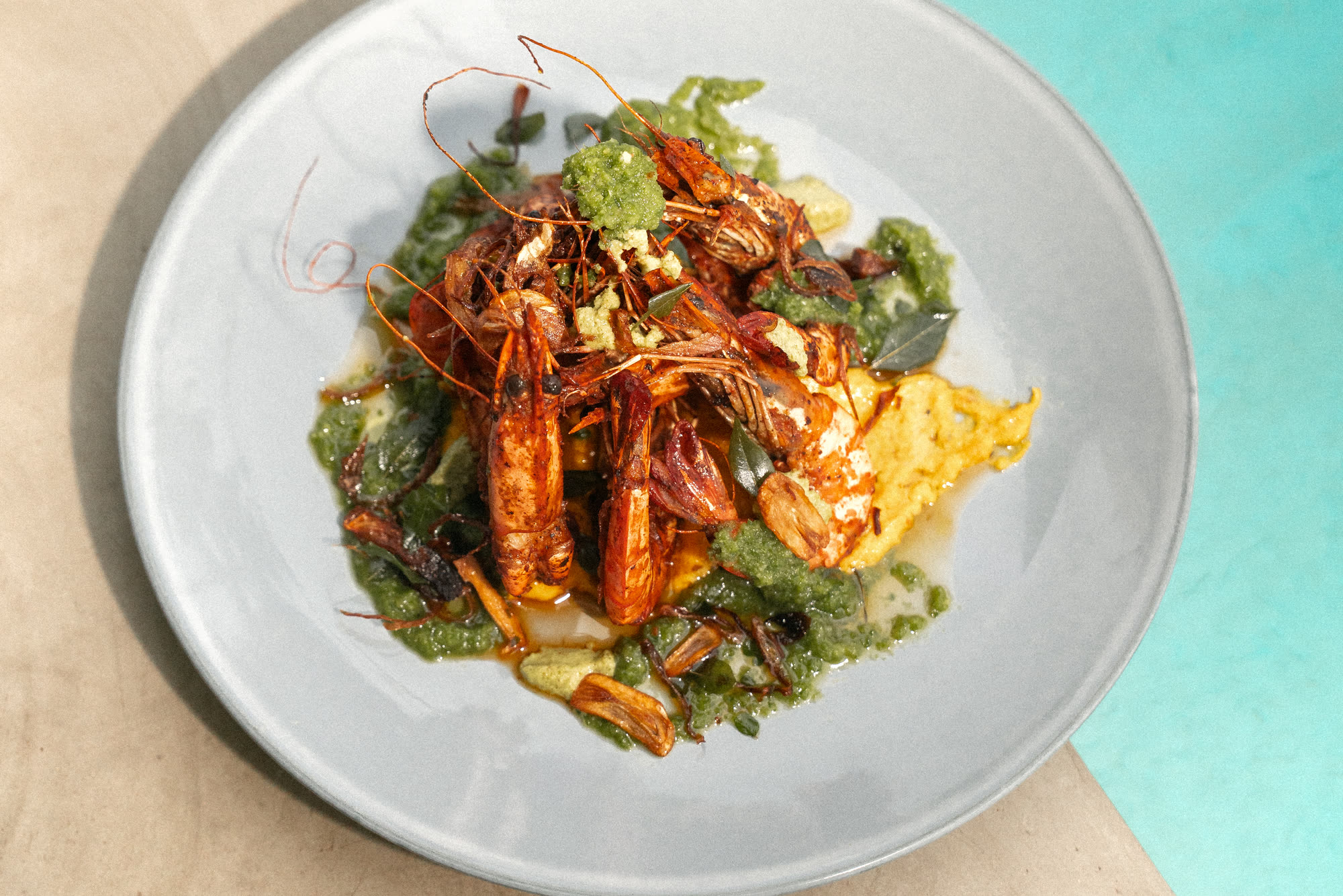 Grilled prawns with a green herb sauce on a grey plate, set on a concrete pool edge beside turquoise water