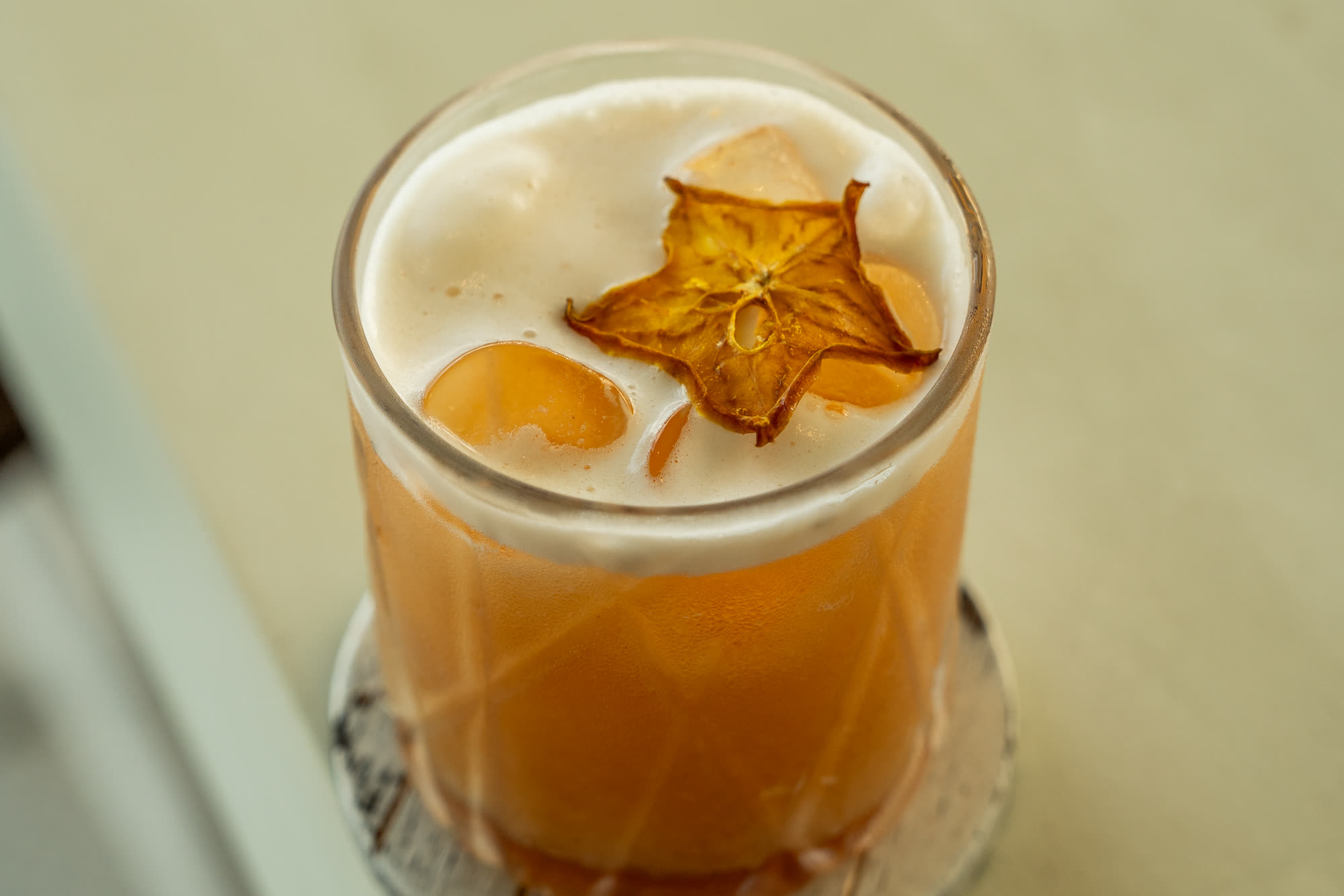 An amber cocktail with a foam top and a slice of dried star fruit, in a cut glass on a whitewashed coaster