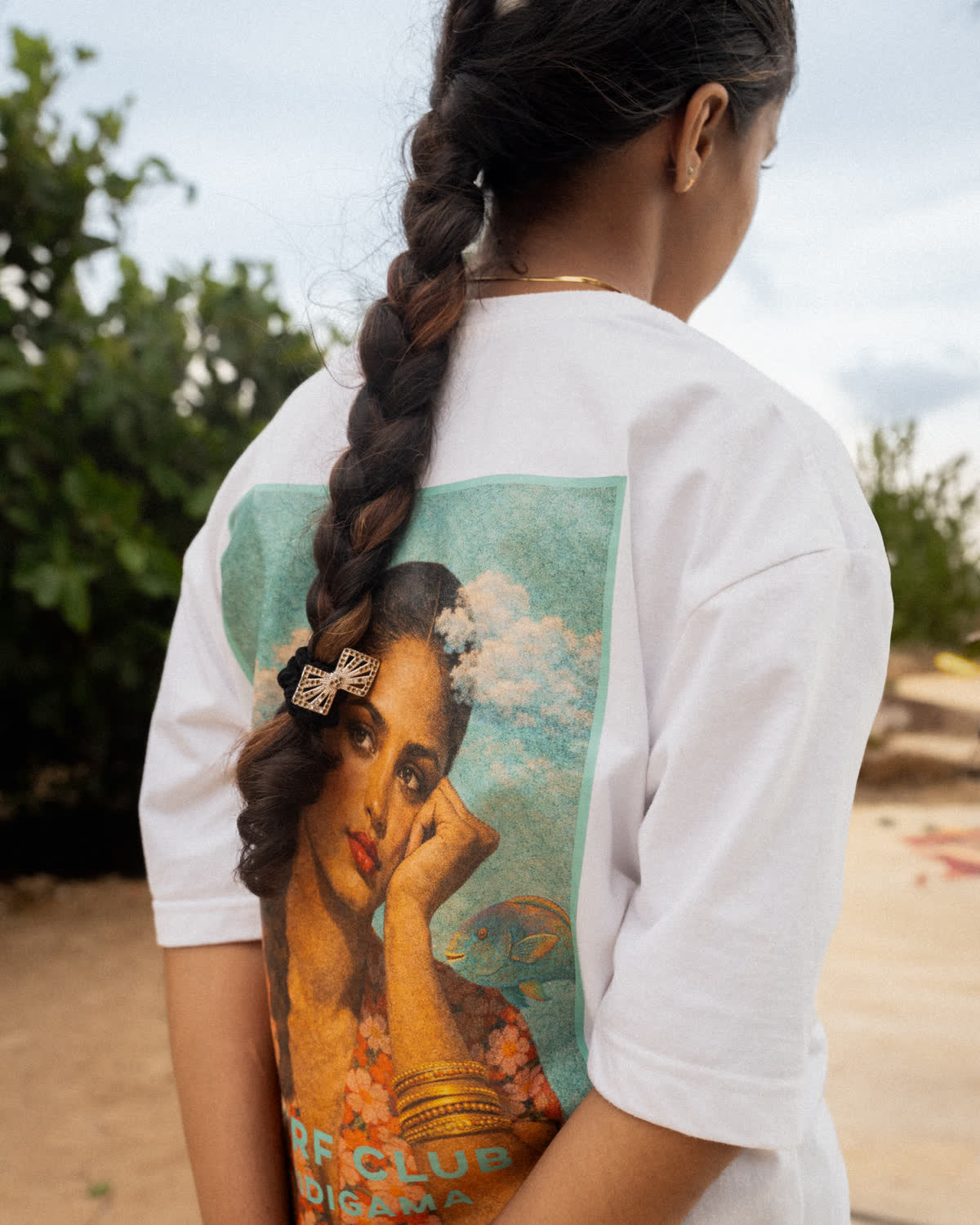 A woman with a long braid seen from behind, wearing a white Surf Club Midigama tee printed with an illustrated portrait, palms and sand behind