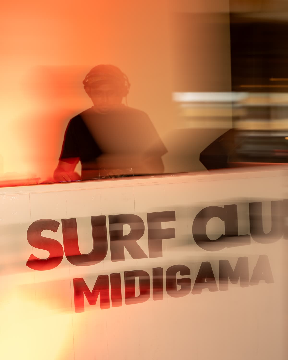 A DJ in headphones behind a white lettered booth, blurred by a slow shutter in orange light
