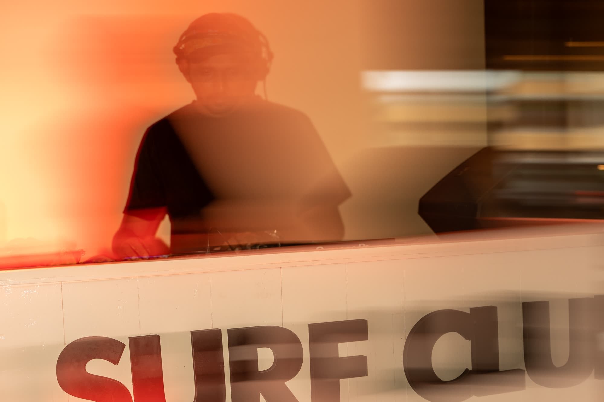 A DJ in headphones behind a white lettered booth, blurred by a slow shutter in orange light