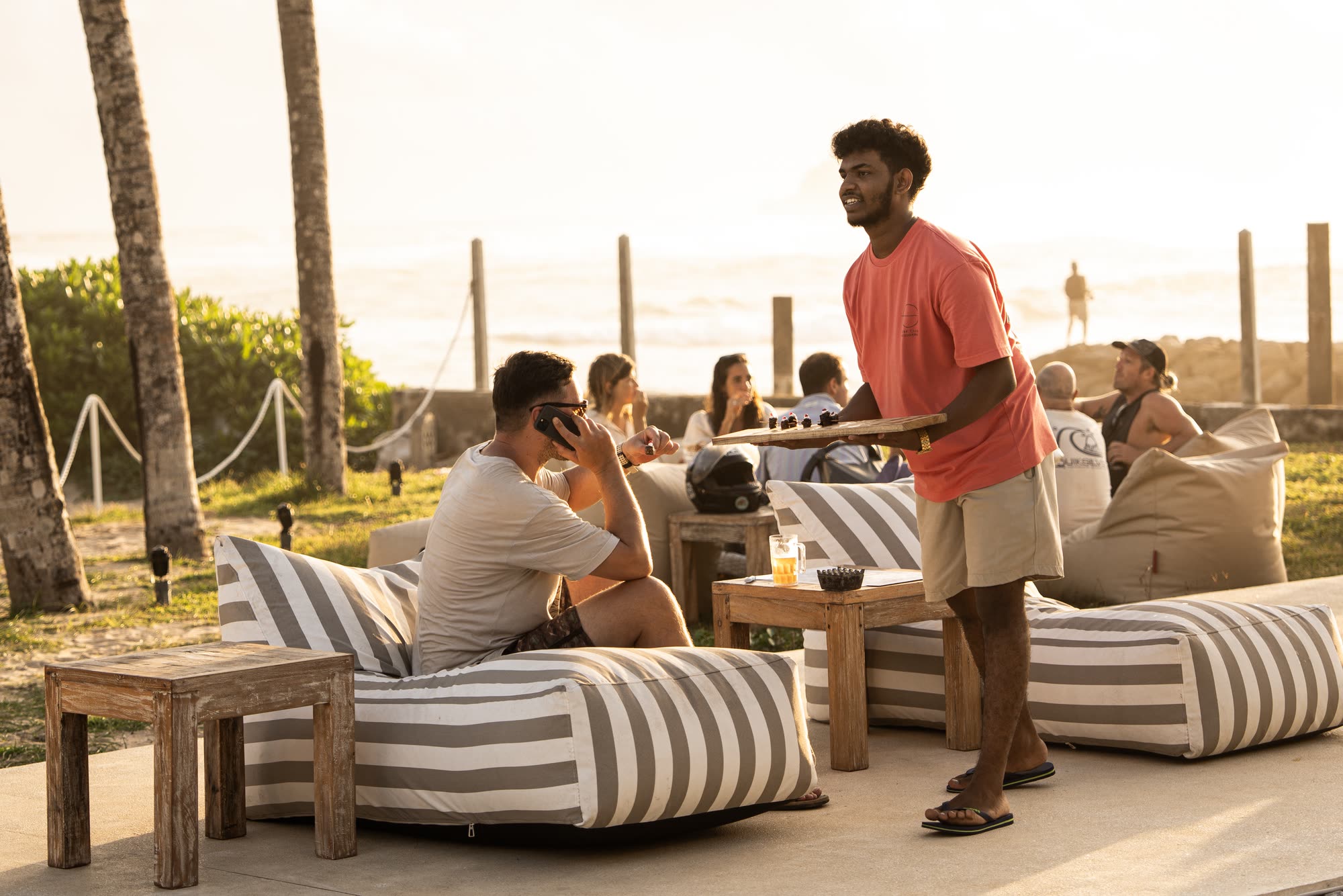 A member of the team carrying a tray past guests on striped bean bags on the terrace at sunset, drinks on timber tables and the lawn, palms and the sea behind