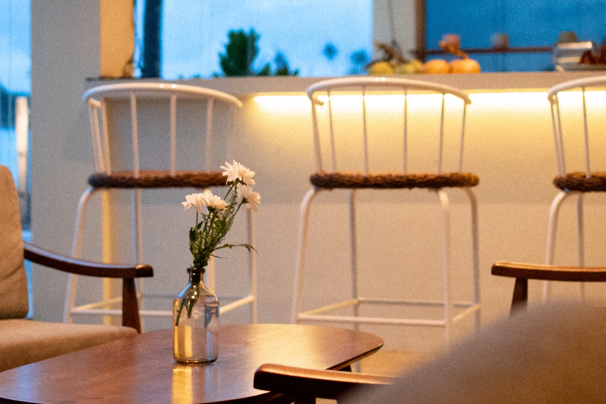 White bar stools with woven seats along a softly lit counter in the evening, white flowers in a glass bottle on a low timber table in front