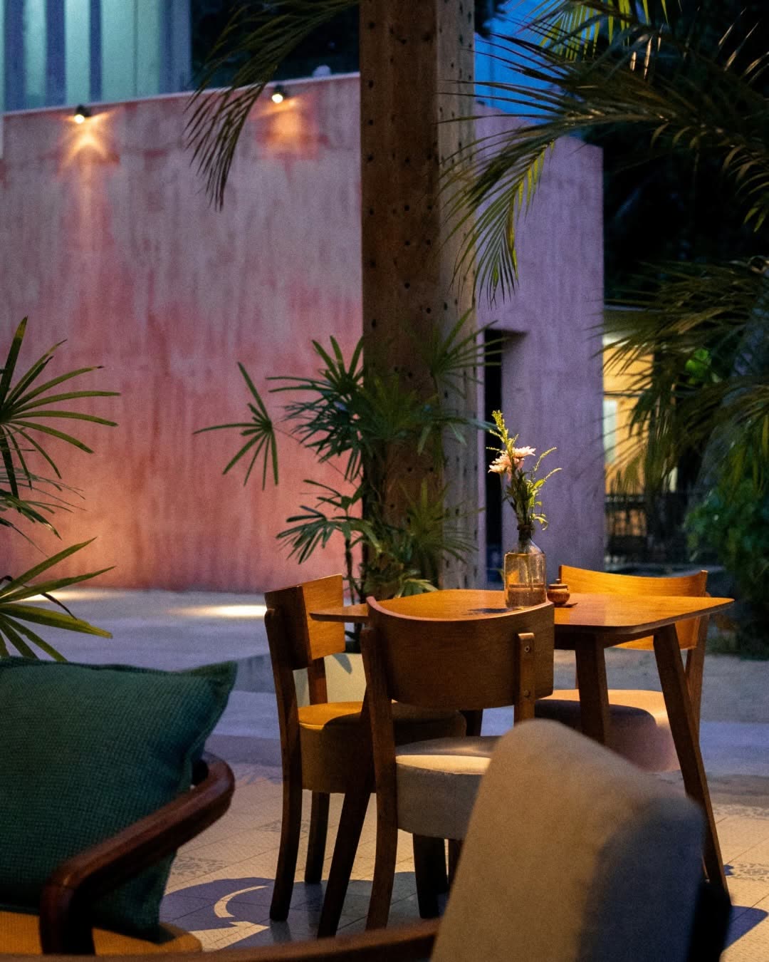 A timber table with a vase of flowers and cushioned chairs in the courtyard at dusk, palms and a pink plaster wall behind