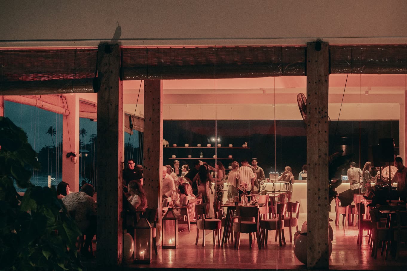 An open-sided dining room at night seen from the garden, glowing pink and full of people at tables and at the bar, candle lanterns at the entrance