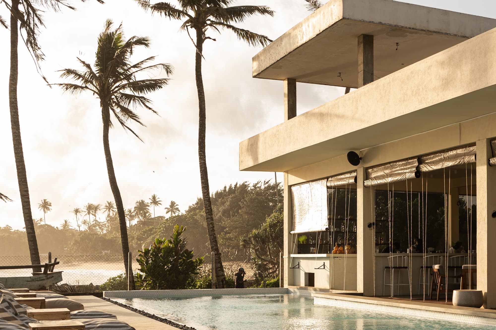 The Club House, a white two-storey building with a glass front, beside the pool in late sun, tall palms and the coast to the left and striped bean bags along the deck