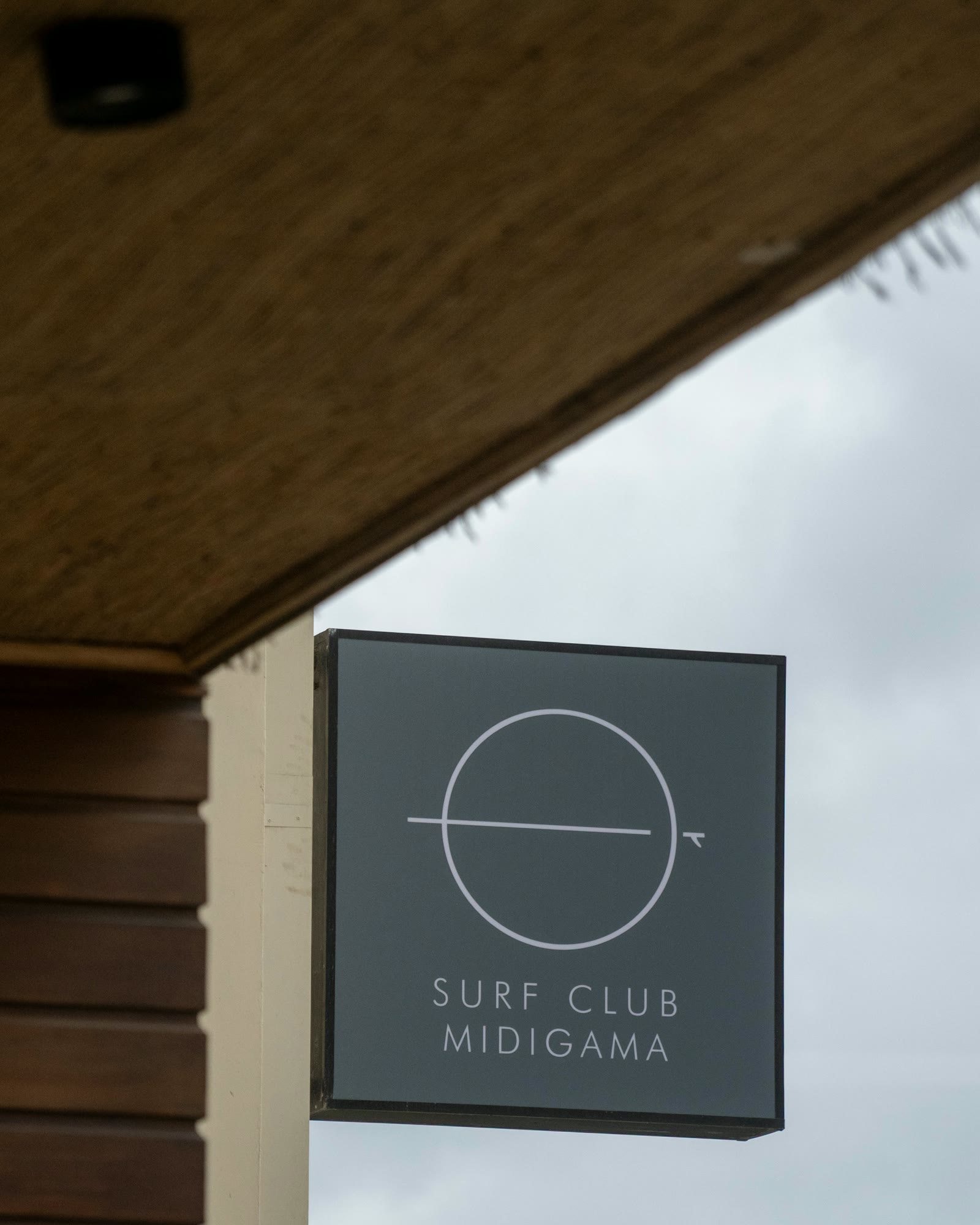 The Surf Club Midigama sign, a dark square lightbox with the circle mark, hanging from a post beside the timber-clad Club House, grey sky behind