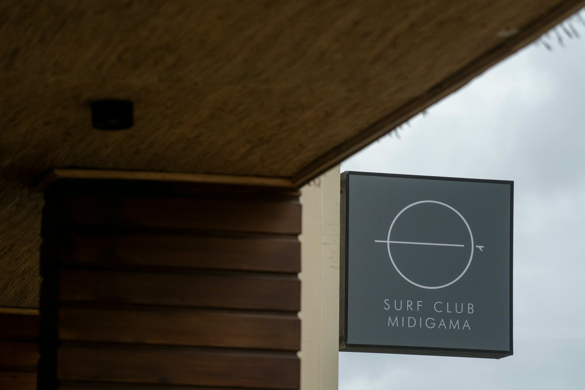 The Surf Club Midigama sign, a dark square lightbox with the circle mark, hanging from a post beside the timber-clad Club House, grey sky behind