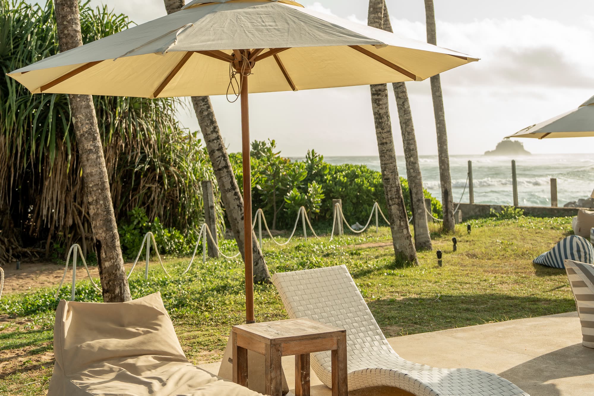 A cream parasol over a white woven lounger, a bean bag and a teak side table at the edge of the terrace, palms and a rope fence behind and a small island on the sea