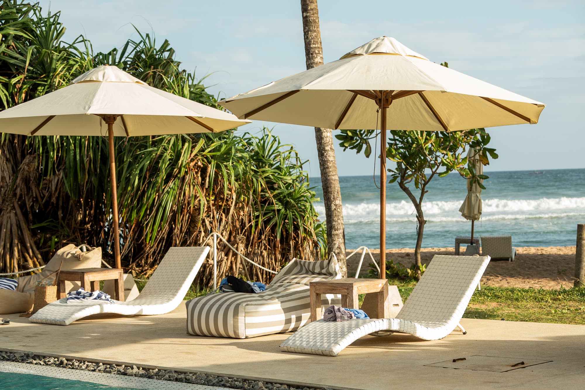 Cream parasols over white woven loungers and a striped bean bag at the edge of the pool deck, pandanus and palms behind and surf breaking on the beach