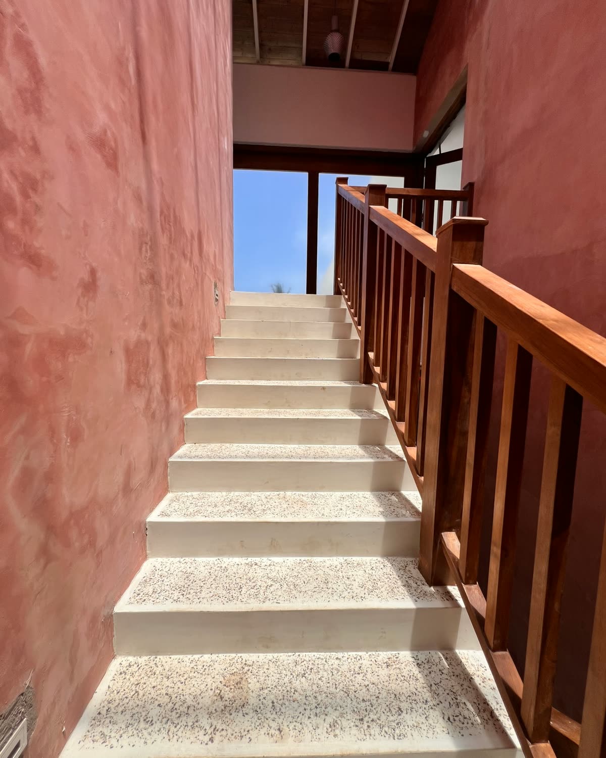 A staircase with white terrazzo steps and a teak handrail between rough pink plaster walls, rising to a timber ceiling and a window of blue sky