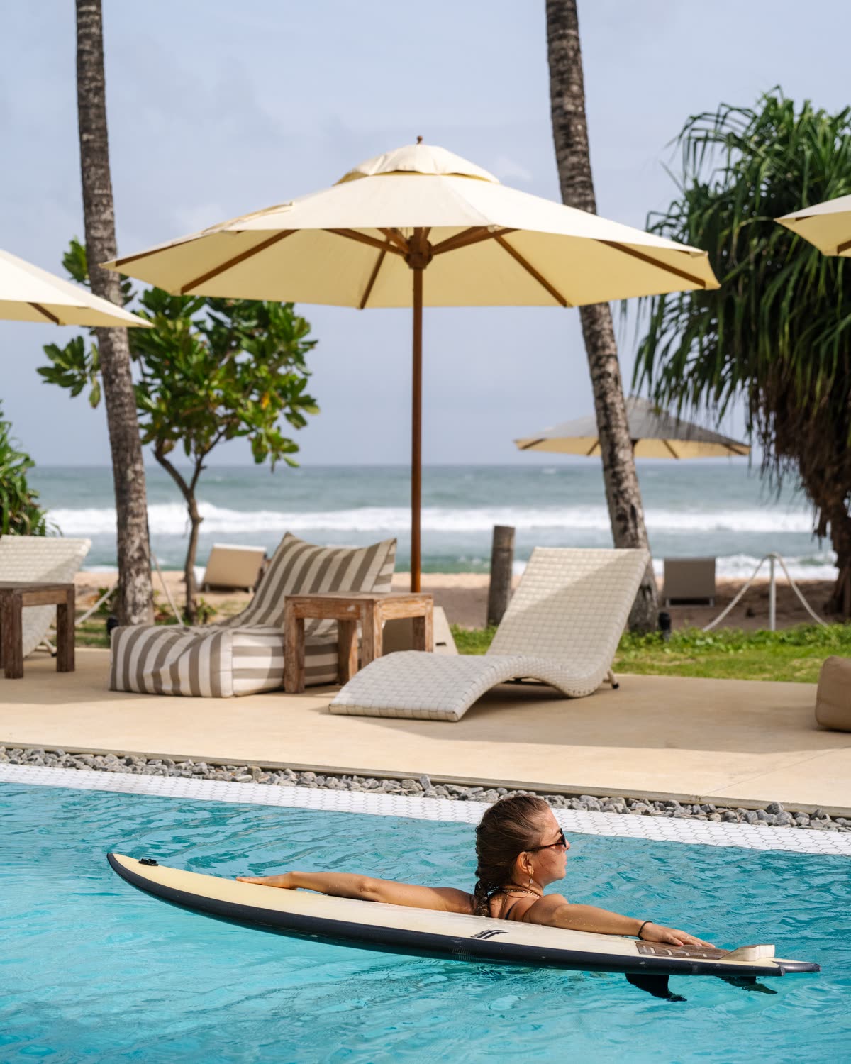 A guest in the pool resting on a white surfboard, a woven lounger, a striped bean bag and cream parasols on the deck behind, palms and surf breaking on the beach