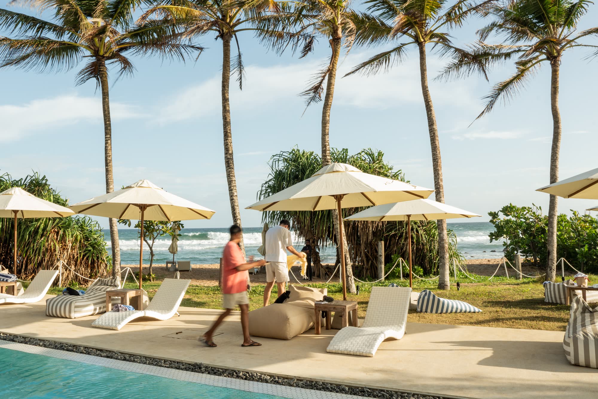 Tall palms over cream parasols, white woven loungers and bean bags on the pool terrace in late afternoon sun, a member of the team walking through and the surf breaking beyond the beach