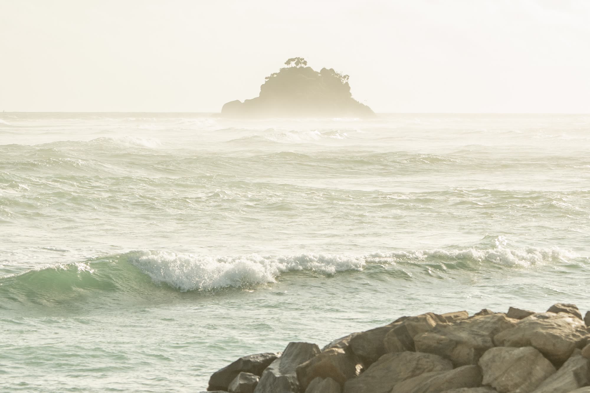 A small tree-topped island on the horizon beyond breaking waves, with rocks in the foreground, in soft hazy light