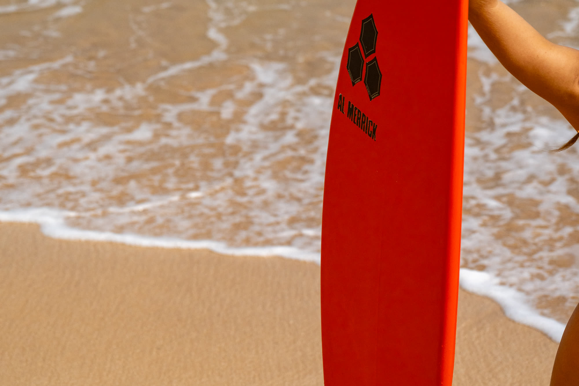 A red surfboard standing upright on wet sand at the water's edge, foam washing in behind