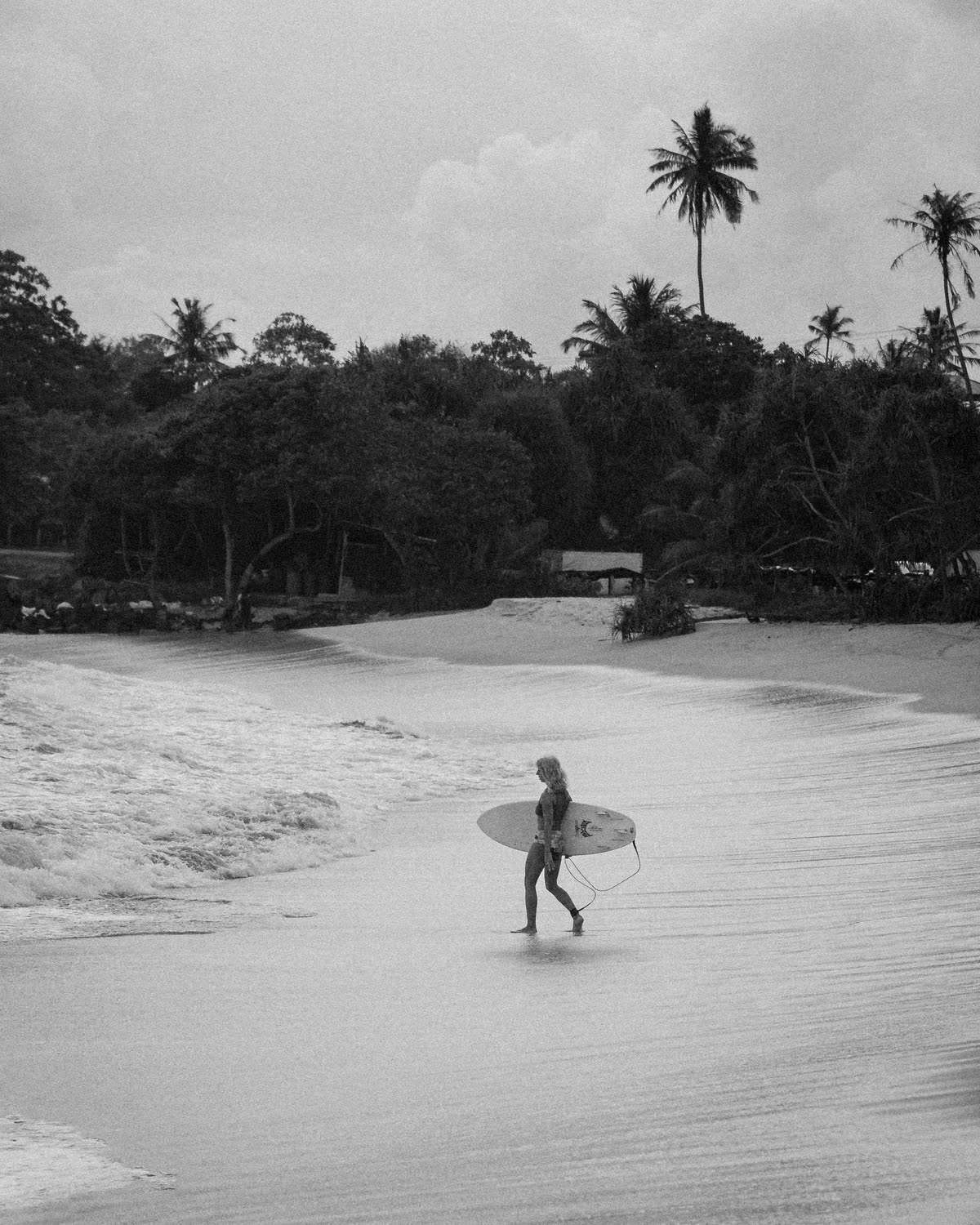 A surfer carrying a board along the shoreline, a wave washing in and palms above the treeline; black and white