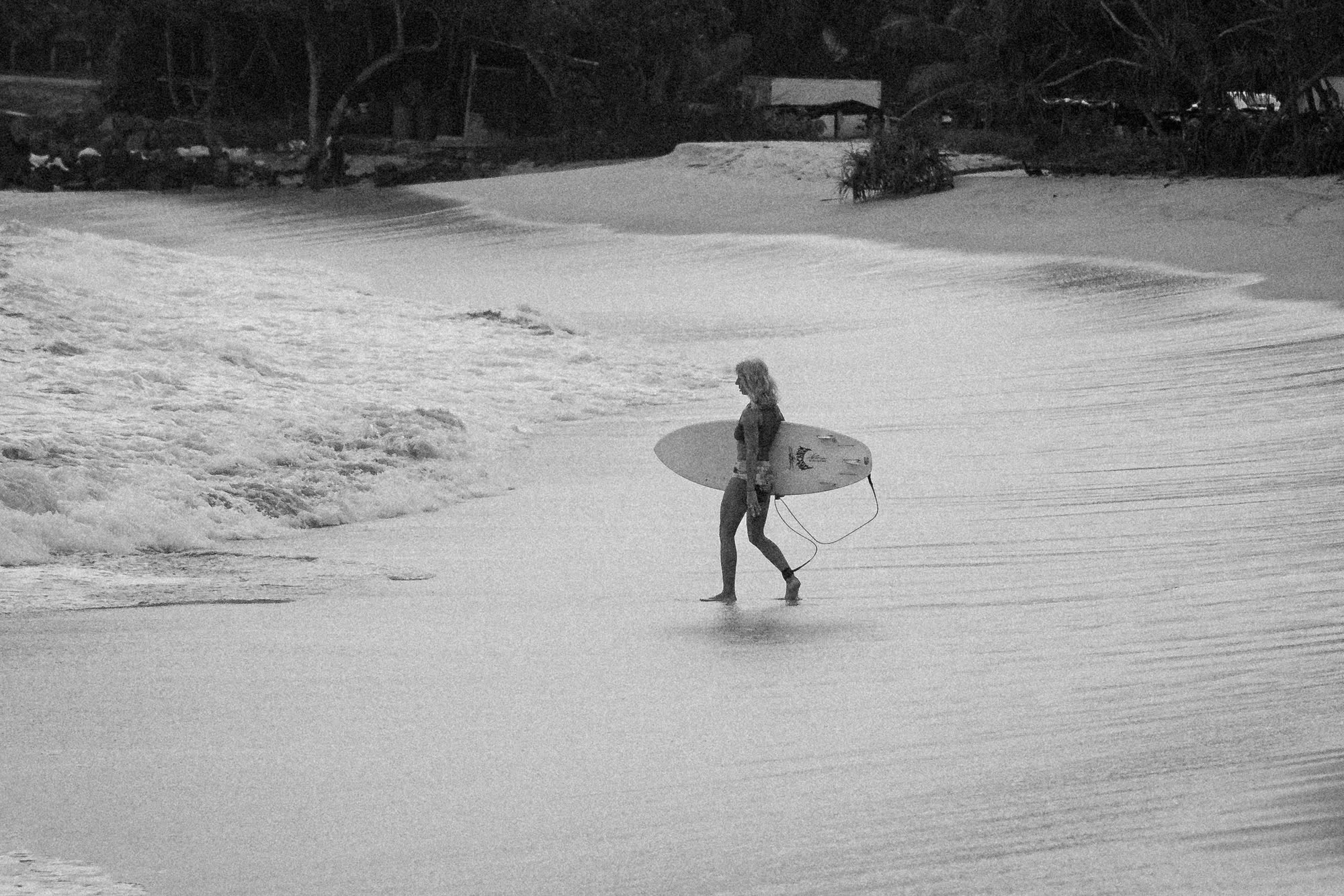 A surfer carrying a board along the shoreline as a wave washes in; black and white